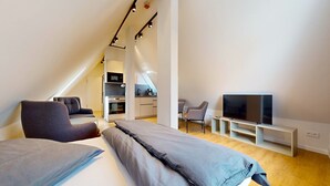 Un dormitorio tipo loft moderno con techo inclinado, una cama grande, un televisor y una kitchenette al fondo.