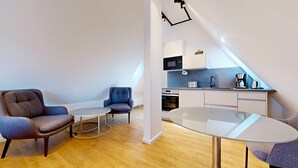 Un apartamento moderno con comedor, dos sillones, una mesita redonda y una kitchenette.