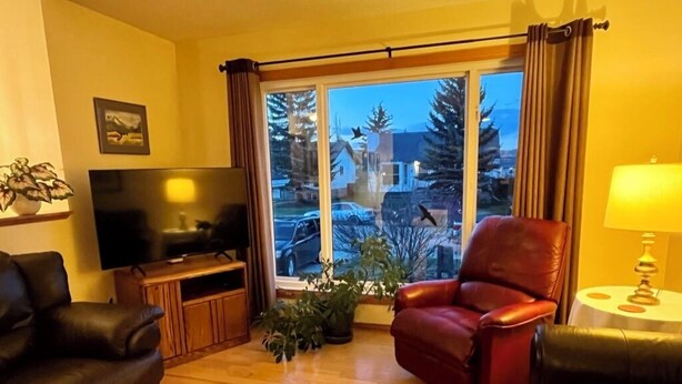 A living room with a television, leather chairs, a small table with a lamp, and a window with curtains. - Bright, spacious 4 bedroom home. (Cochrane)