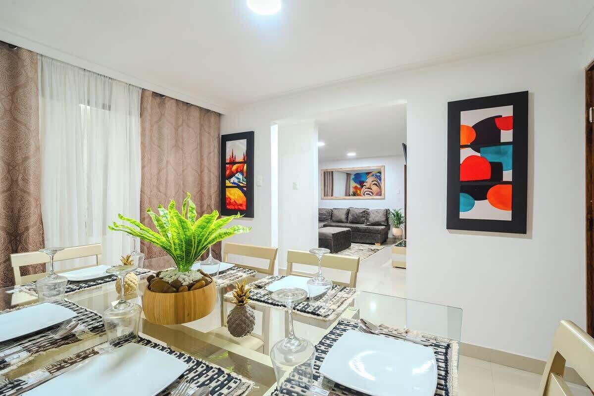 A dining area with a glass table, white plates, and a potted plant. There are abstract paintings on the walls and a sofa in the background.