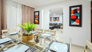Un comedor con una mesa de vidrio, platos blancos y una planta en maceta. Hay pinturas abstractas en las paredes y un sofá al fondo.