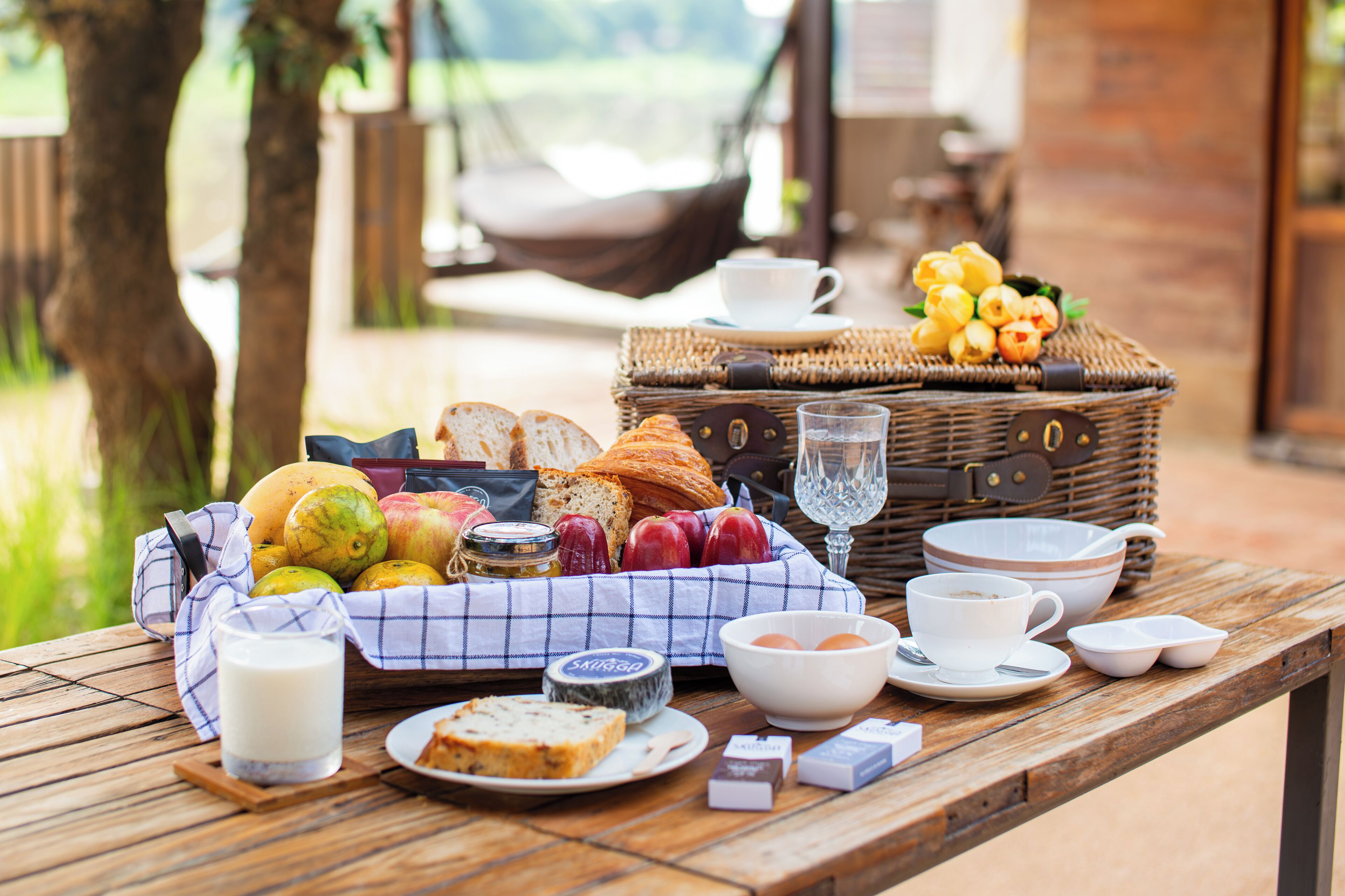 Desayuno con pan, frutas, queso y bebidas sobre una mesa de madera al aire libre.