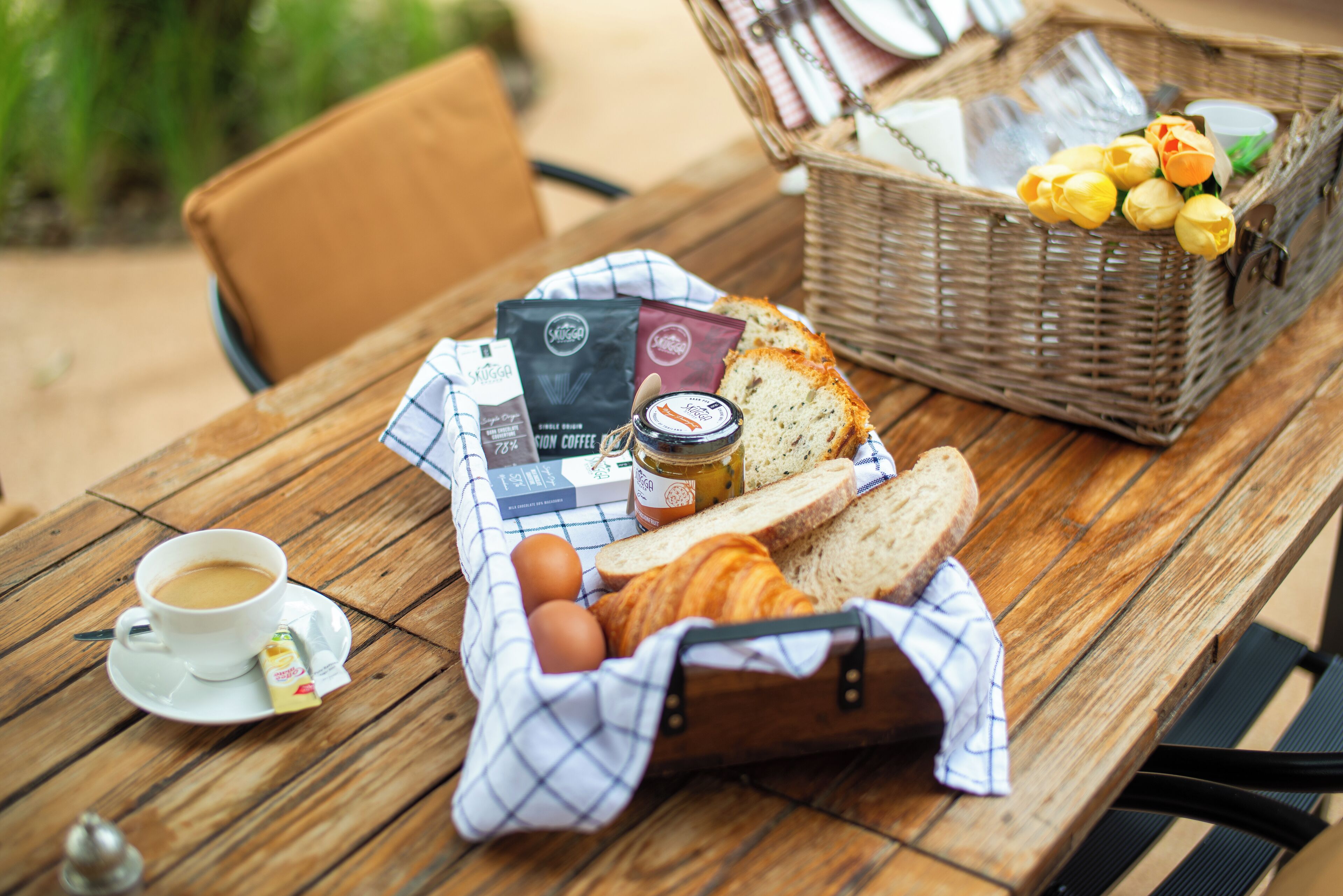 Ett träbord med en kopp kaffe, bröd, ägg och en korg som innehåller blommor och ett glas med sylt.