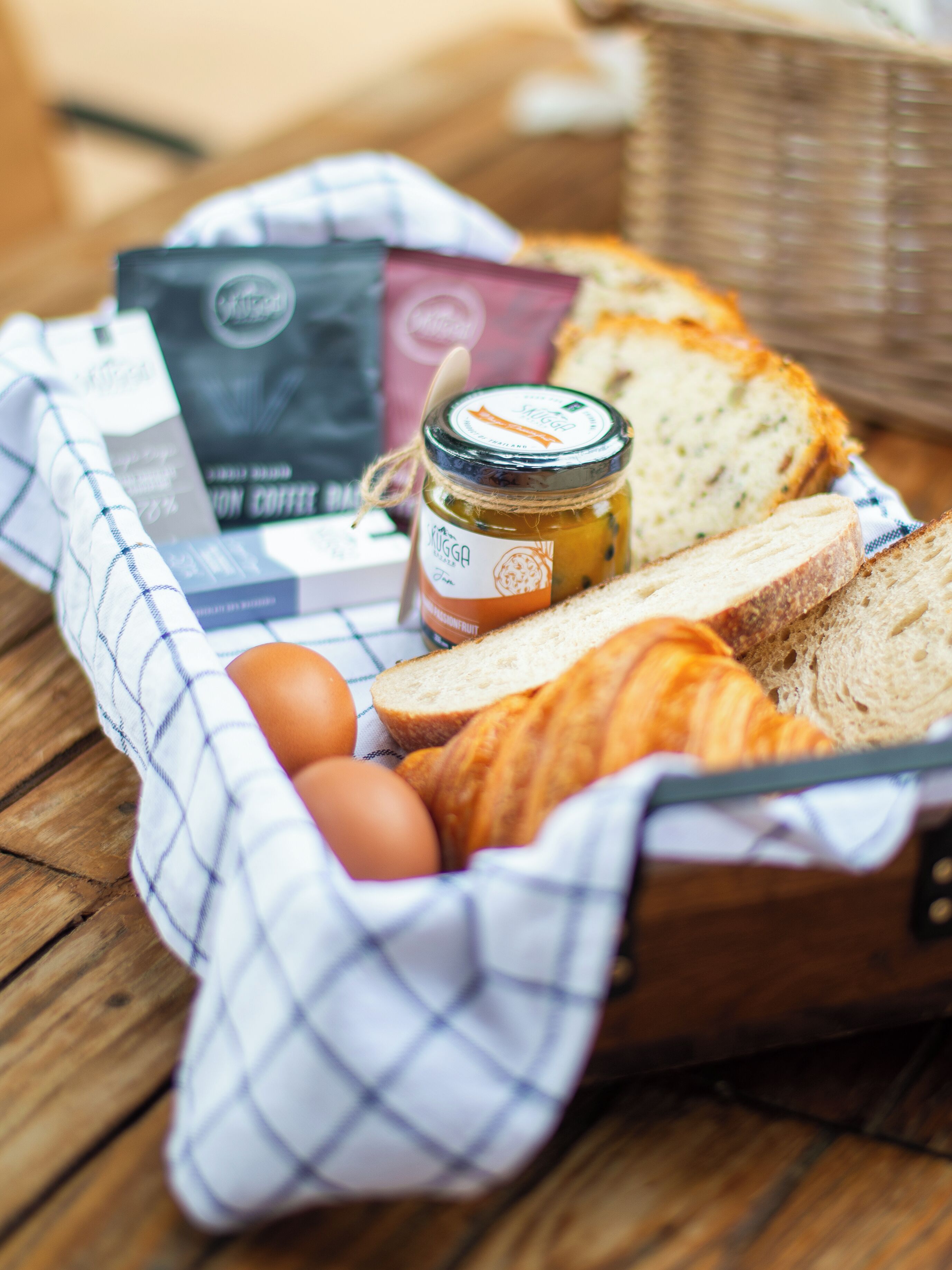 A basket containing bread, eggs, a jar of preserves, and coffee packets on a wooden table.
