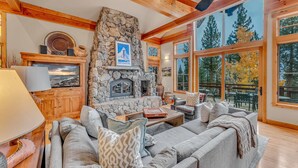 A cozy living room with a stone fireplace, wooden beams, and large windows offering a view of trees. - Northstar Ski-In/Ski-Out with Hot Tub & Views (Northstar)