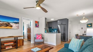 A modern living room with a wooden coffee table, a blue sofa, and a kitchen area with grey cabinets and a stainless steel refrigerator.