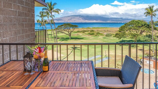 A balcony with a wooden table, wicker chair, and a view of the ocean and mountains.