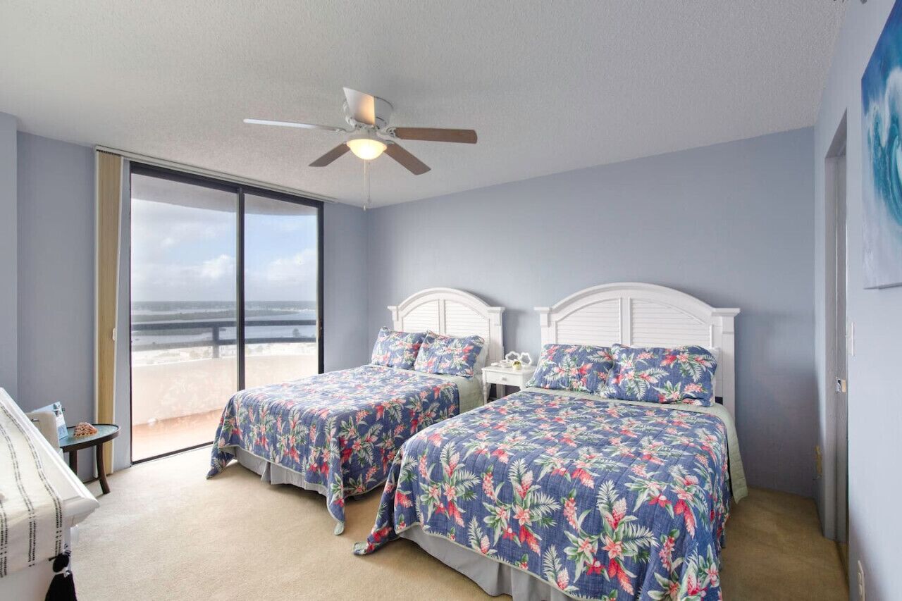 Ein Schlafzimmer mit zwei Betten, einem Deckenventilator und Blick auf den Strand durch ein großes Fenster.
