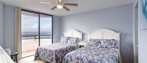 Ein Schlafzimmer mit zwei Betten, einem Deckenventilator und Blick auf den Strand durch ein großes Fenster.