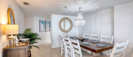 Un comedor con mesa de madera, sillas blancas, una araña y un espejo decorativo.