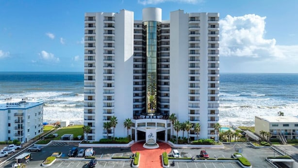 A high-rise hotel with a central glass elevator shaft, a red circular driveway, and a view of the ocean.