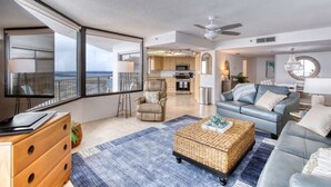 A spacious living area with a large window, a ceiling fan, a kitchen in the background, and a grey sofa.