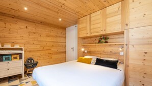A wooden-paneled bedroom with a bed, bedside table, and a small shelf with decorative items. - Epinettes : Vue montagne et déco contemporaine. (LE GRAND BORNAND)