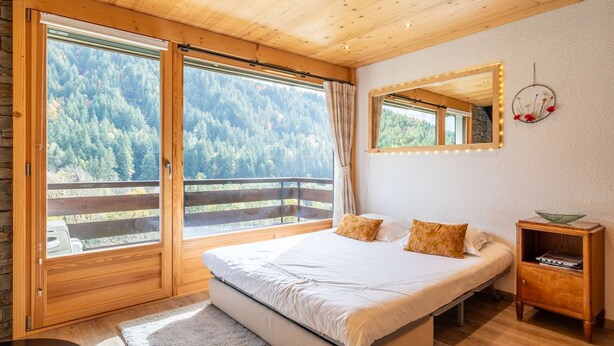 A bedroom with a large bed, wooden floor, a nightstand, a mirror, and a view of a forested area through the windows. - Epinettes : Vue montagne et déco contemporaine. (LE GRAND BORNAND)