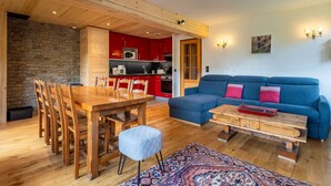 A modern living space with a wooden dining table, blue sofa, and a kitchen in the background. - Réséda : Calme Montagnard avec Vue Apaisante (LE GRAND BORNAND)