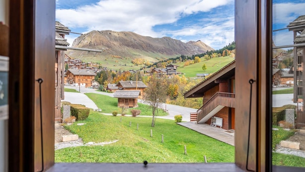 A view from a window of a building with a mountain in the background, a grassy area, and several wooden buildings. - Réséda : Calme Montagnard avec Vue Apaisante (LE GRAND BORNAND)