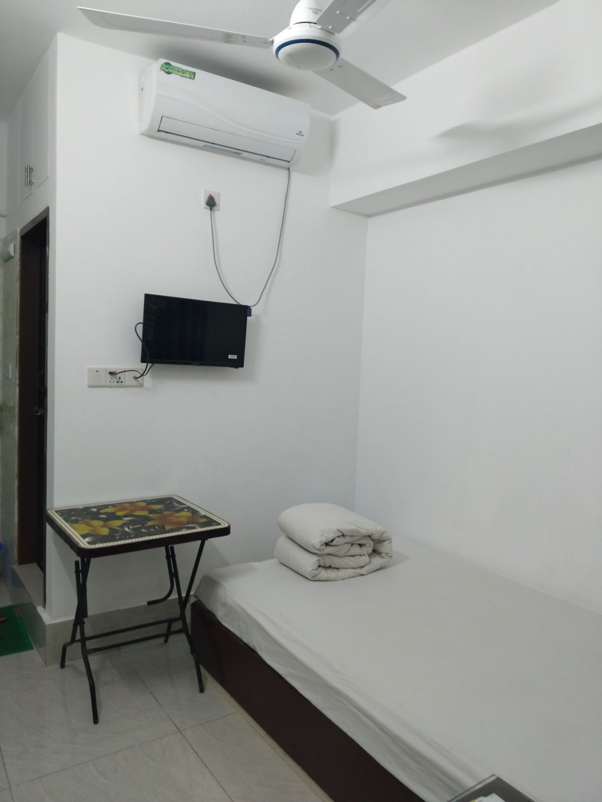 Una habitación pequeña y limpia con cama, mesa, aire acondicionado y ventilador de techo.