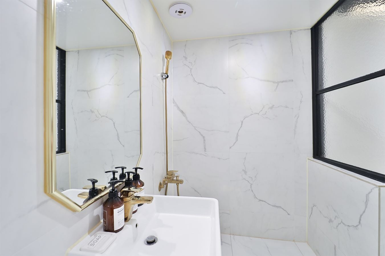 Un baño con pared de mármol blanco, espejo con marco dorado, lavamanos blanco y ducha con cabezal de ducha dorado.