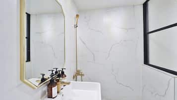 A bathroom with a white marble wall, a gold-framed mirror, a white sink, and a shower with a gold showerhead.