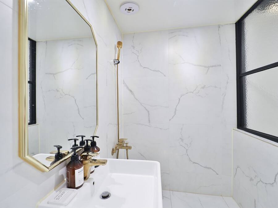 A bathroom with a white marble wall, a gold-framed mirror, a white sink, and a shower with a gold showerhead.
