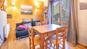 A cozy room with a wooden floor, a sofa with cushions, a wooden dining table with chairs, a bookshelf, and a window with a view of trees. - Nice apt 4 pers near the slopes (Chamrousse)