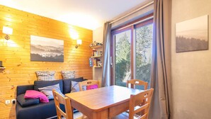 A cozy room with a wooden wall, a sofa with cushions, a table with chairs, a bookshelf, and a large window with curtains. - Nice apt 4 pers near the slopes (Chamrousse)
