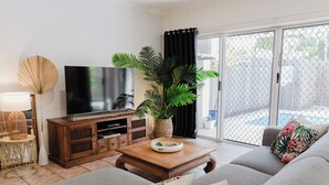 Una sala de estar con un sofá, una mesa de centro, un televisor sobre un mueble de madera, una planta en maceta y vistas a través de un ventanal.