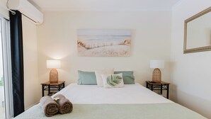 Habitación de hotel con una cama, dos mesitas de noche con lámparas, un cuadro con motivos playeros en la pared y una ventana con cortinas.