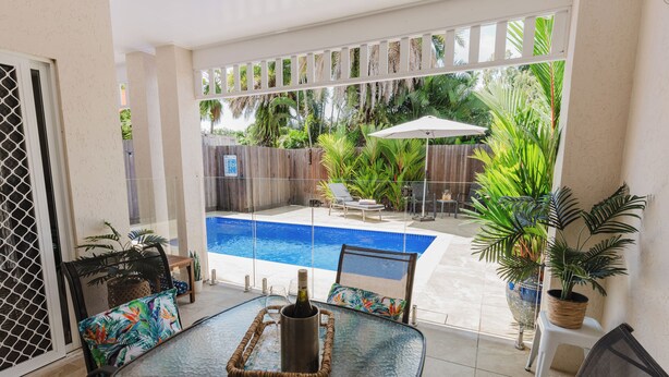 Una mesa con superficie de vidrio, con una botella de champán, rodeada de sillas con cojines estampados con motivos tropicales, con vista a un jardín trasero con piscina y abundante vegetación.