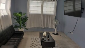 A living room with a black sofa, a coffee table, a potted plant, a TV, and a standing fan. - Charming family friendly home,furnished with parking facilities,near malls/beach (Nassau)