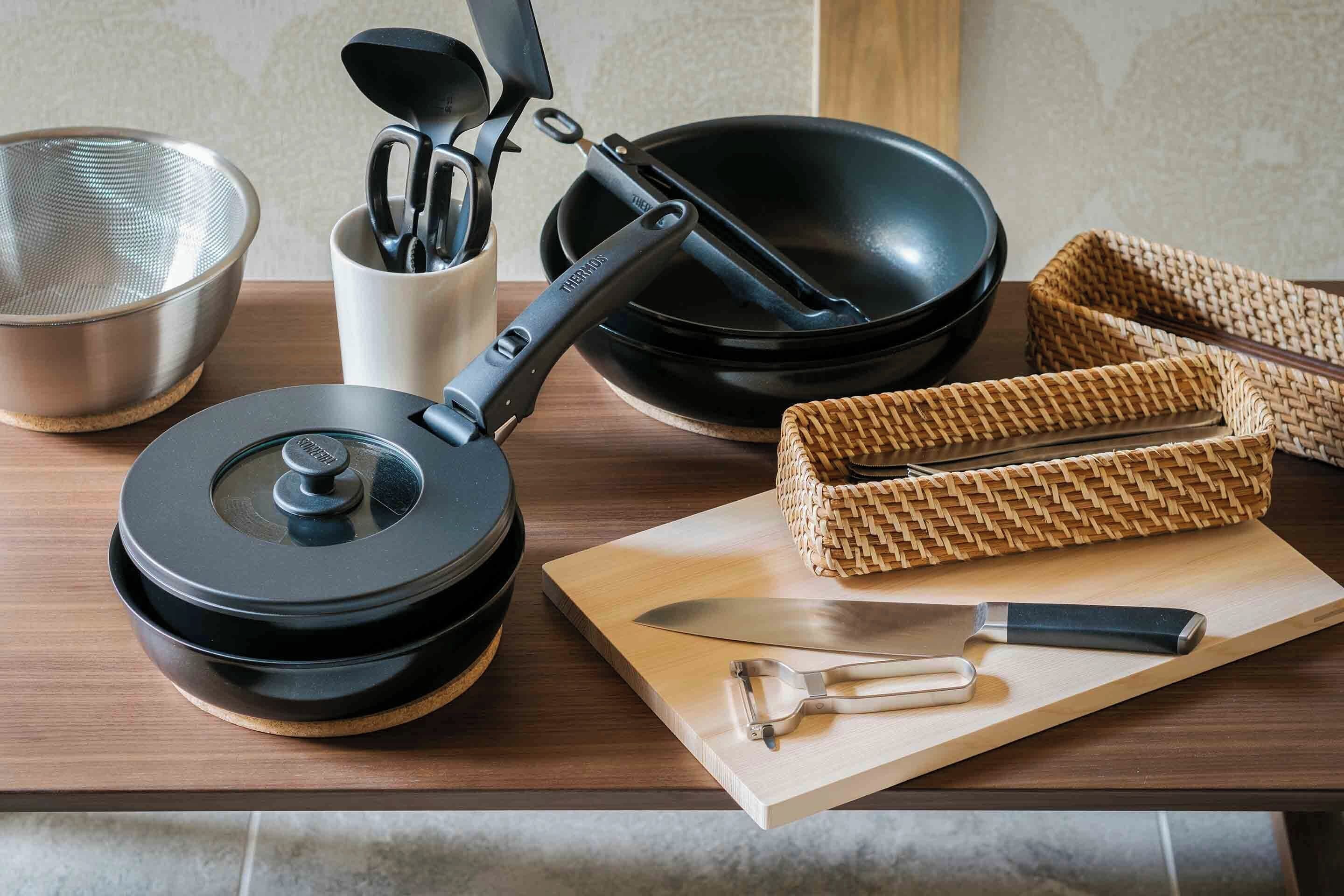 Un plan de travail de cuisine avec un cuiseur à riz électrique noir, une poêle noire, un panier en osier, un couteau et une passoire en métal.