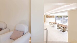 Interior moderno com sofá branco, sala de jantar com mesa e cadeiras de madeira, e vista através de grandes janelas.