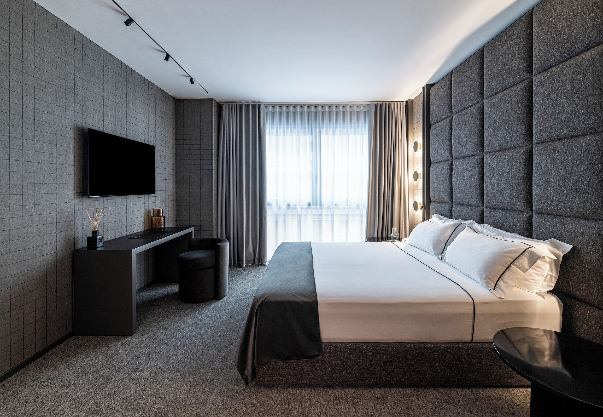 Une chambre d’hôtel moderne dotée d’un grand lit, d’un téléviseur à écran plat fixé au mur, d’un bureau avec une chaise et offrant une vue sur la ville grâce à de grandes fenêtres.