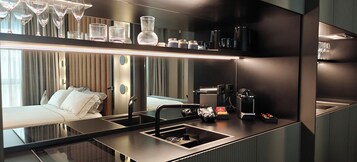A modern hotel room with a large bed, a built-in kitchen with a sink, and a minibar.