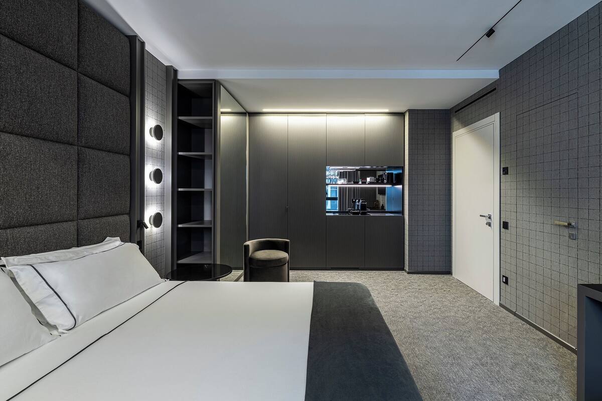 A modern bedroom with a large bed, a seating area, a vanity with a mirror, and a kitchenette in the background.