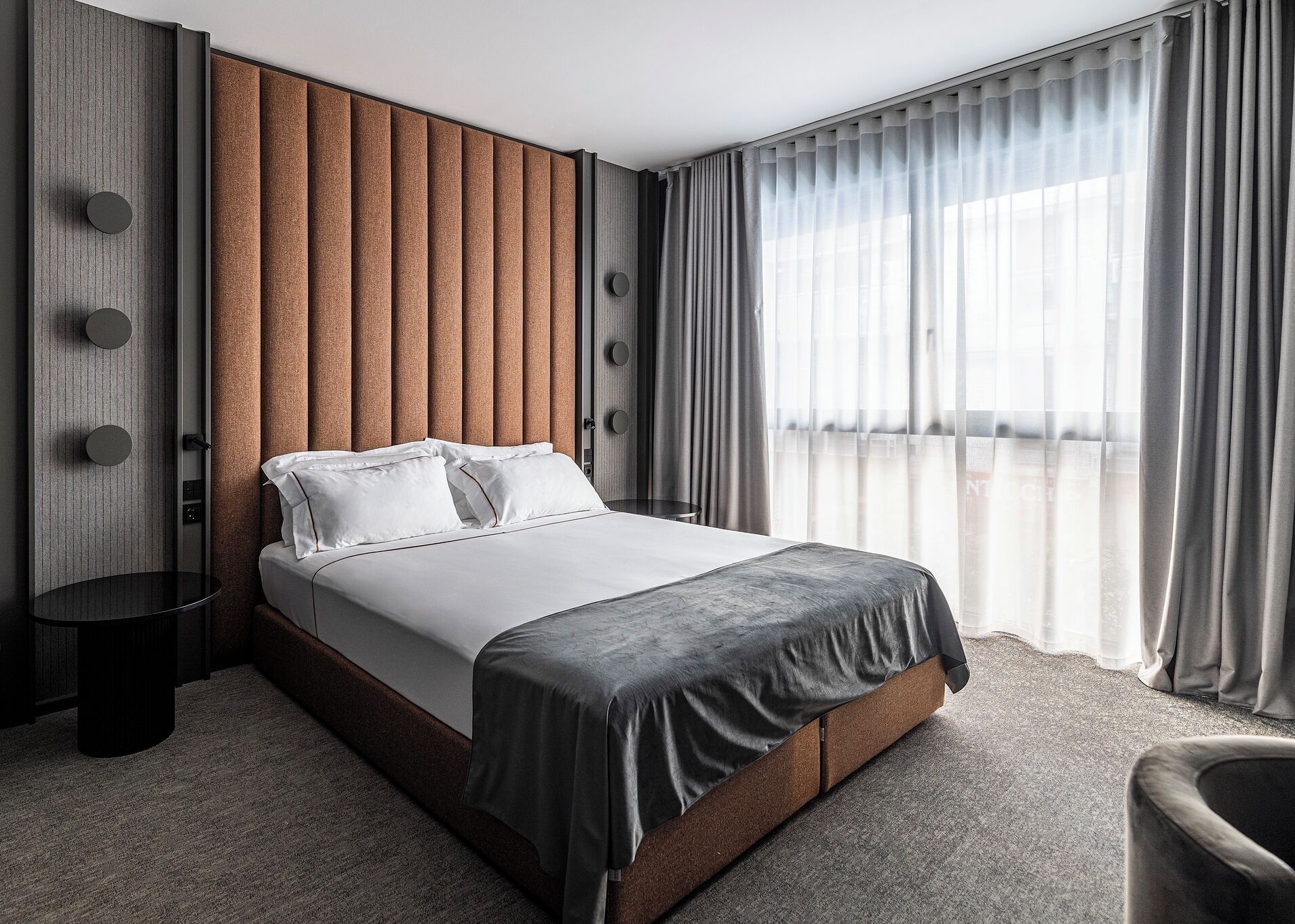 Una moderna camera d'hotel con un ampio letto, una testiera in legno, un tavolino rotondo e vista sulla città attraverso tende trasparenti.