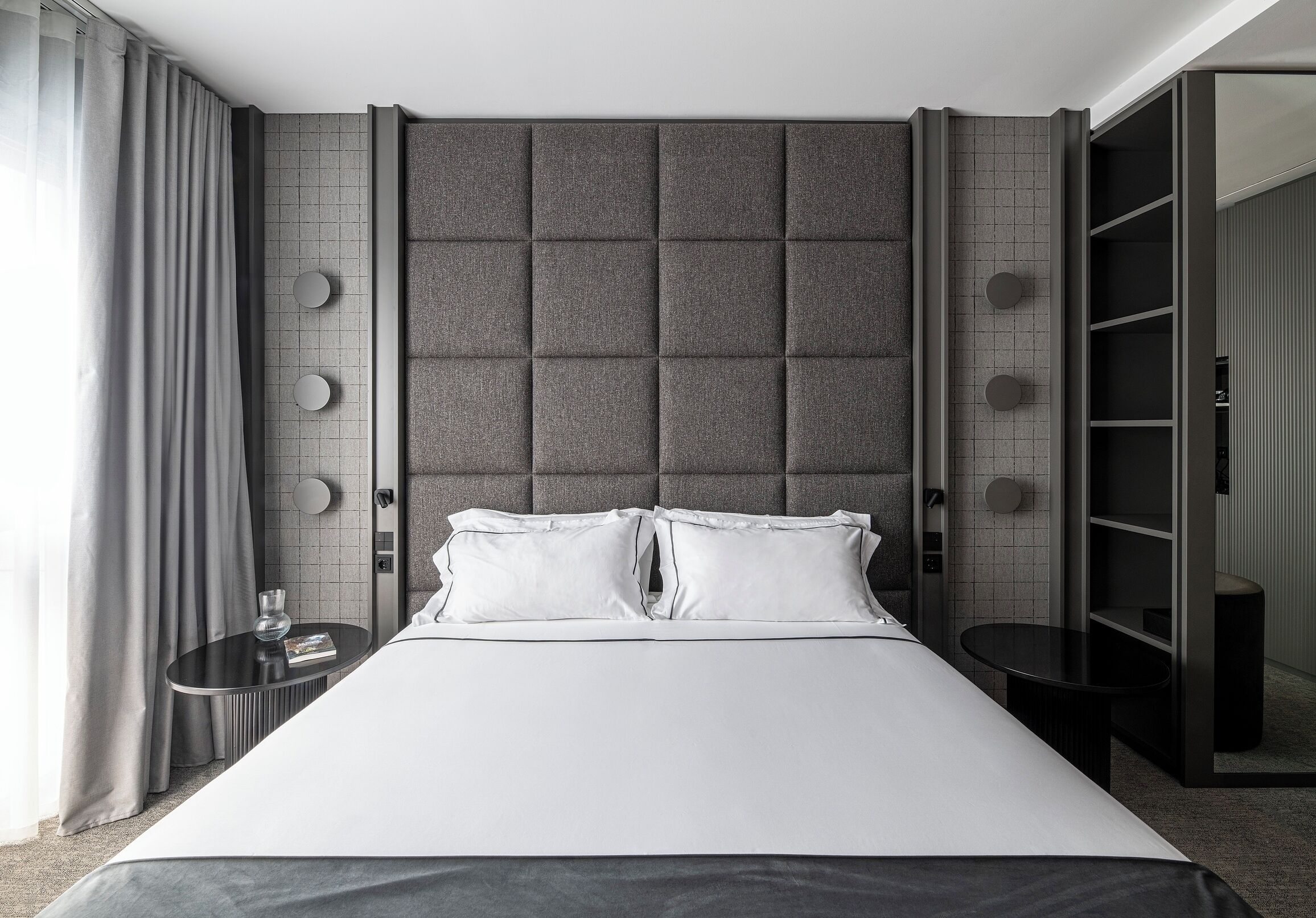 Ein modernes Hotelzimmer mit einem großen Bett, einem gepolsterten grauen Kopfteil, einem runden Nachttisch mit einem Glas und einer eingebauten Garderobe mit Regalen.