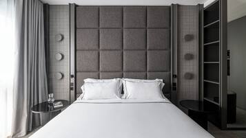 Ein modernes Hotelzimmer mit einem großen Bett, einem gepolsterten grauen Kopfteil, einem runden Nachttisch mit einem Glas und einer eingebauten Garderobe mit Regalen.
