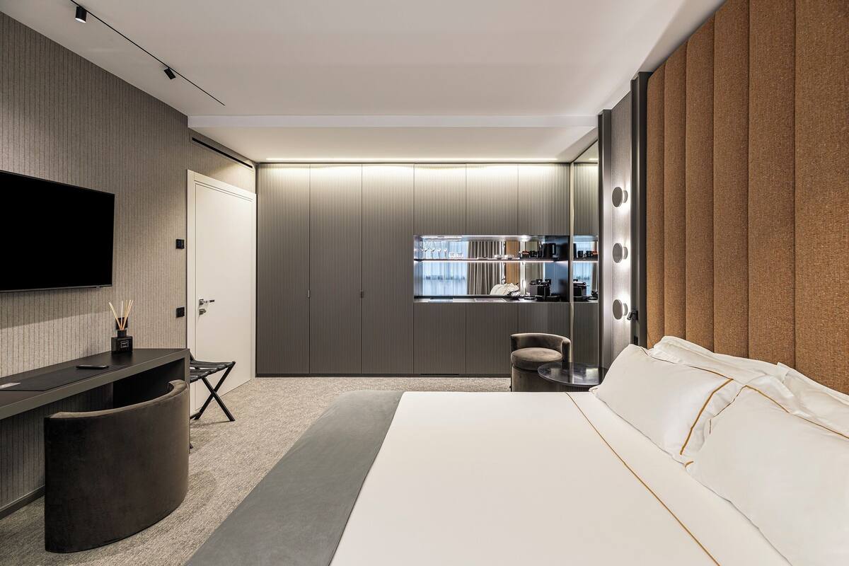 A modern hotel room with a large bed, a desk with a flat-screen TV, a seating area, and a kitchenette.