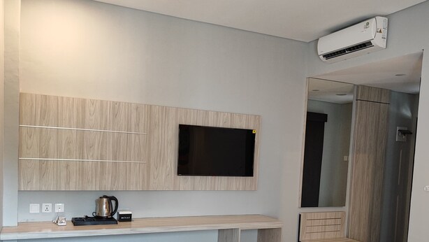 Een moderne hotelkamer met een bed, een televisie bevestigd aan een houten paneel, een klein bureau met een waterkoker en een prullenbak.