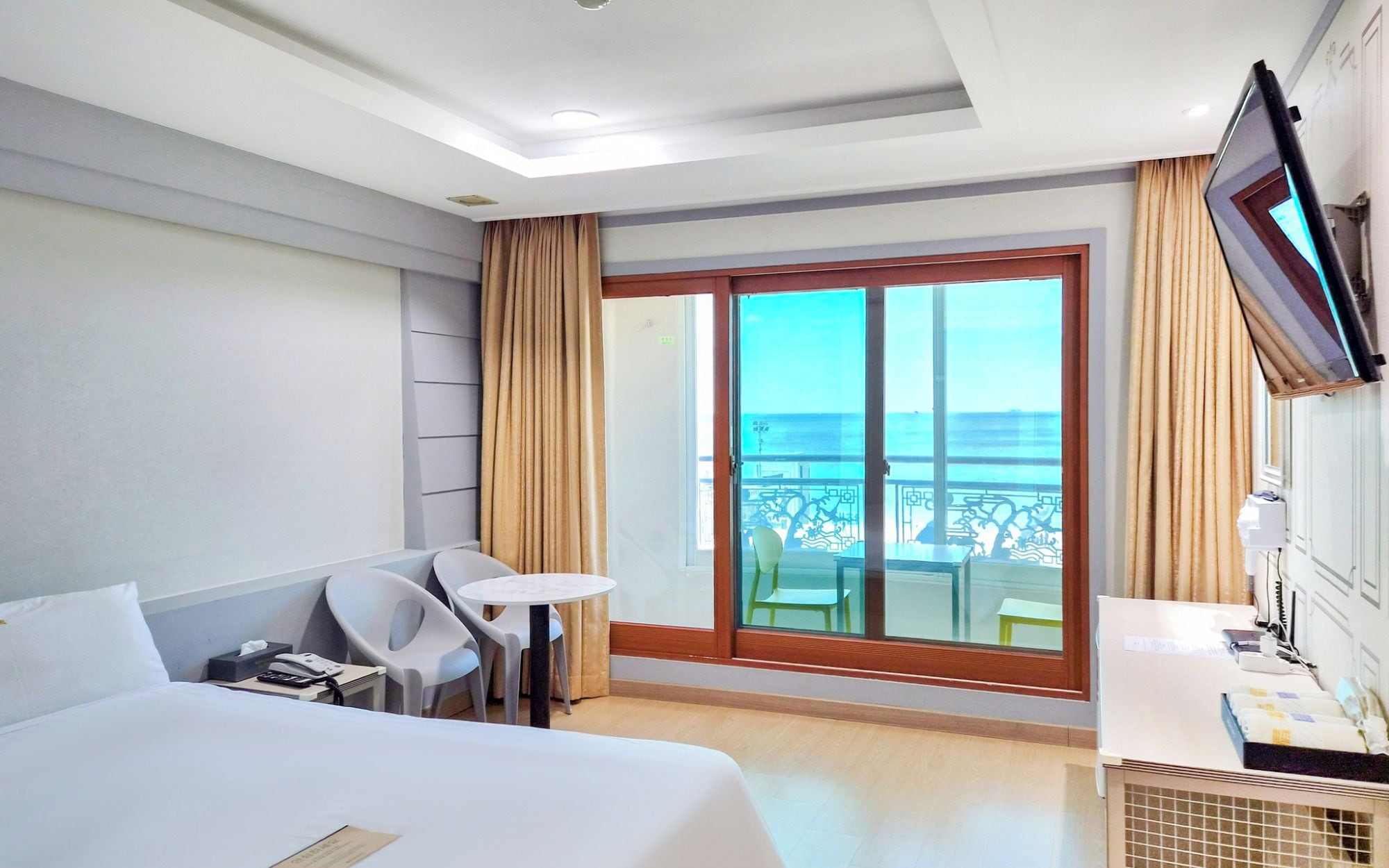 Habitación de hotel con cama, escritorio, silla, mesita, televisor y balcón con vista al mar.