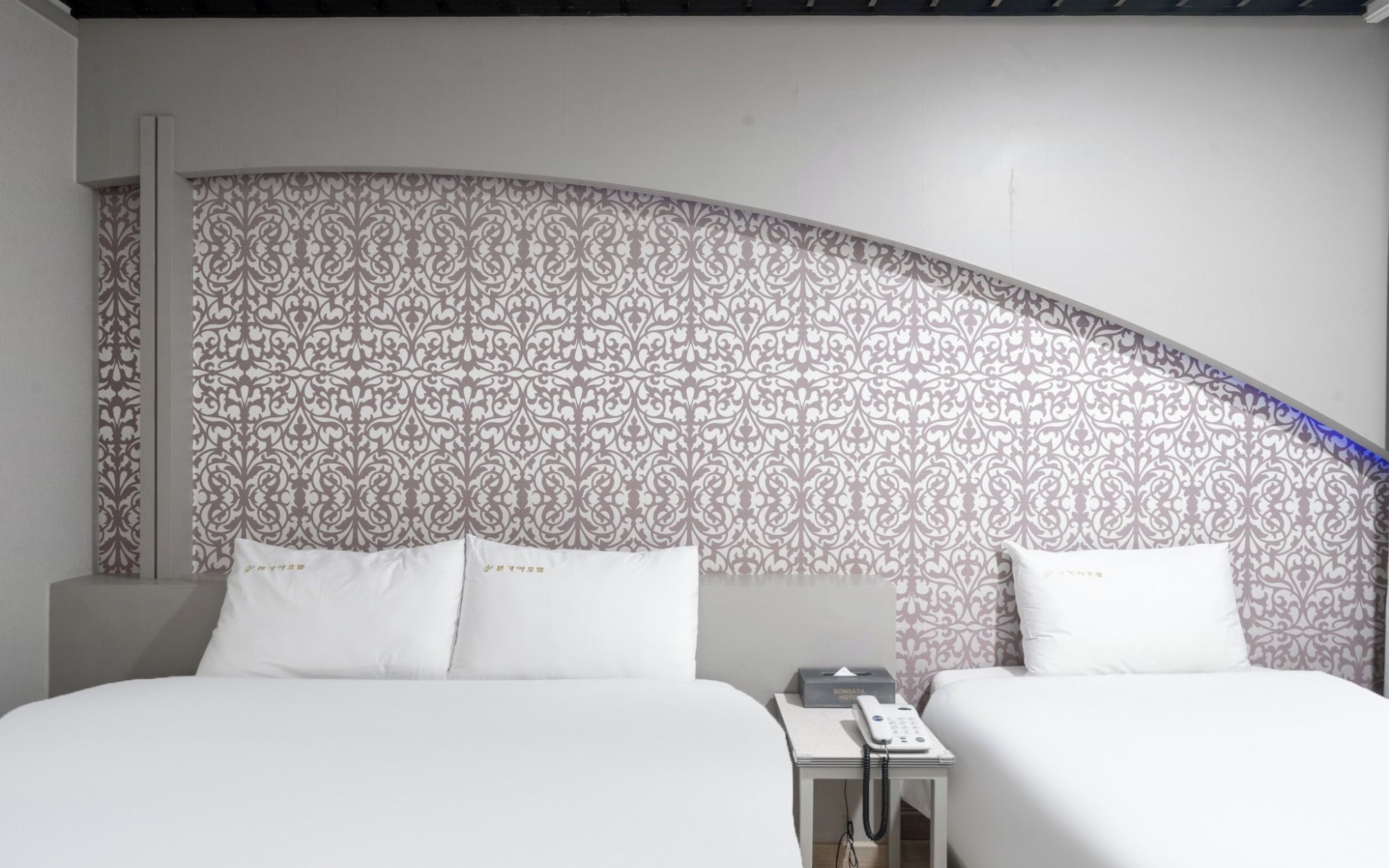 Habitación de hotel con dos camas, una pared curva con papel tapiz estampado y una mesita de noche con un teléfono.