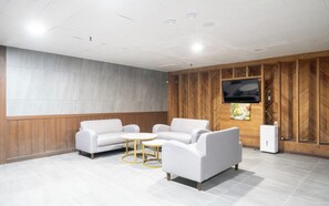 Un espace d’attente moderne avec un téléviseur à écran plat fixé à un mur en bois, deux canapés blancs, une petite table ronde et un radiateur blanc.