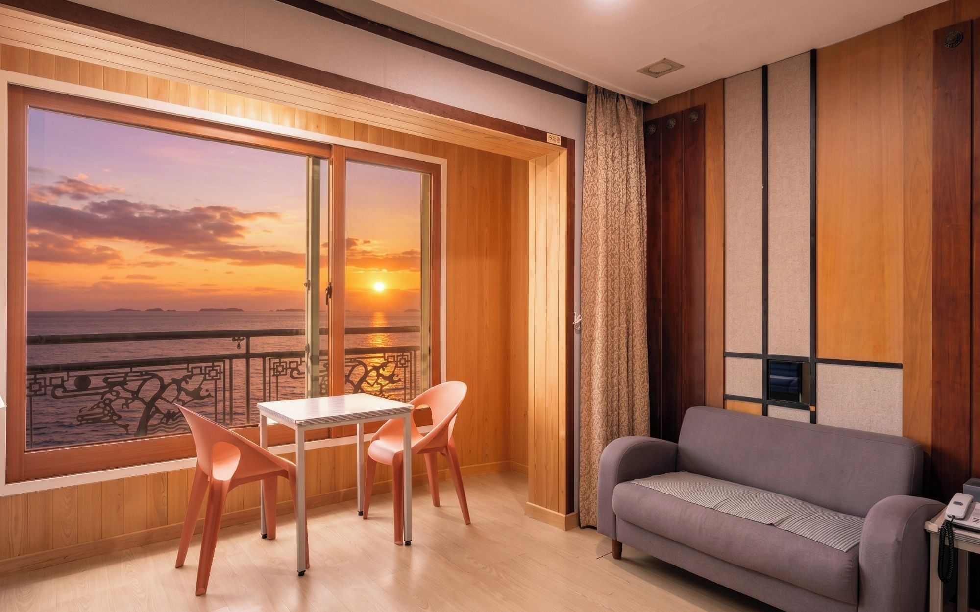 Una habitación con vista a una puesta de sol sobre el agua, un sofá, una mesa con sillas y una cortina.