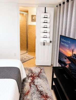 Une chambre d’hôtel avec un lit, un téléviseur diffusant une image de paysage, une porte sur laquelle sont accrochées des serviettes, et une vue sur une plage et un phare.