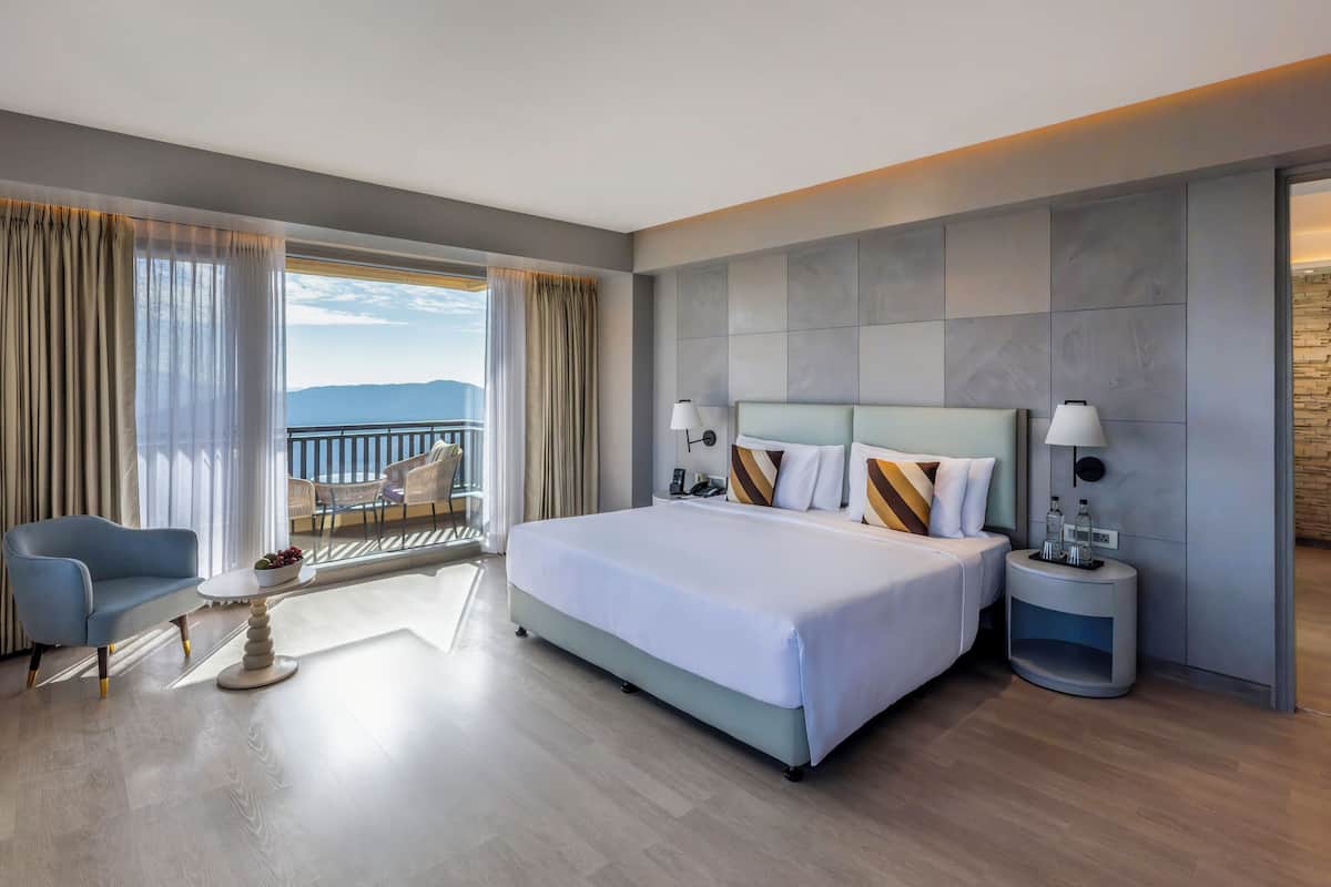 A hotel room with a large bed, a sitting area with a chair and a table, a nightstand with a lamp, and a view of the outdoors.