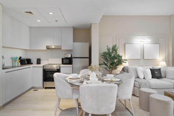 A modern kitchen with white cabinets, a dining area with a glass table and chairs, and a living area with a sofa and ottoman. - Maison Privee - Exquisite Living with Scenic Canal Vws on Yas Island (Abu Dhabi)