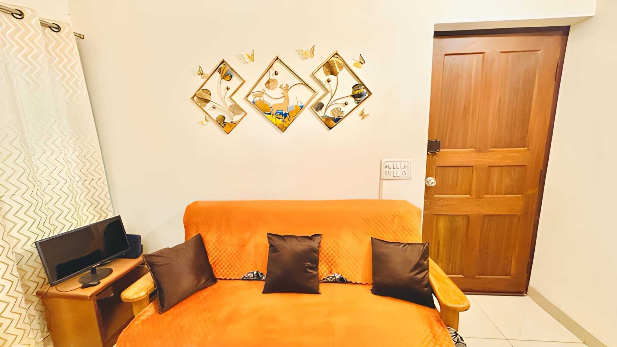 Un dormitorio con una cama naranja, dos almohadas, una mesita con un ordenador portátil y una puerta de madera.