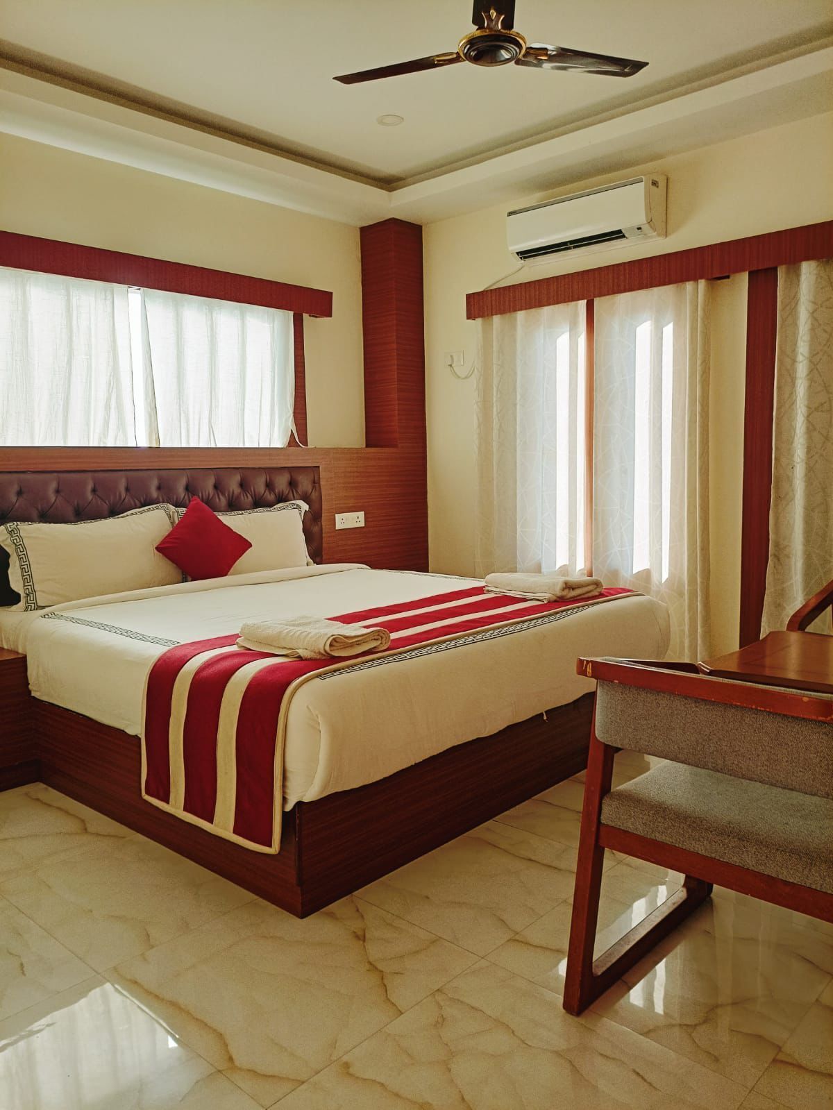 Quarto de hotel com uma cama grande, cabeceira de madeira, ventilador de teto, ar condicionado e uma pequena mesa de madeira.