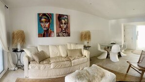 Una sala de estar con un sofá color beige, sillas blancas, una mesa de comedor y dos cuadros en la pared.